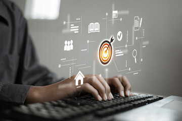 A person types on a keyboard while various digital business icons such as a target, documents, and users float in mid-air, representing goals and data management.