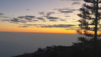 A serene sunset view over the horizon, with soft clouds and silhouettes of trees and houses framed against a vibrant orange sky.