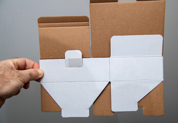 A hand holds a flat, unfolded cardboard box with brown and white surfaces, showing pre-cut flaps, folds, and a glued tab, ready to be assembled for packaging use.