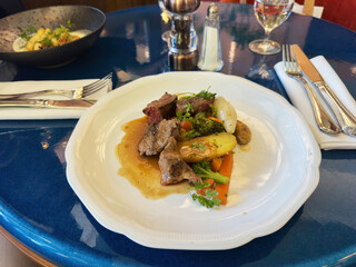 A gourmet French main course featuring tender slices of meat, glazed vegetables, and savory jus on a white plate set on a glossy blue table in a fine restaurant