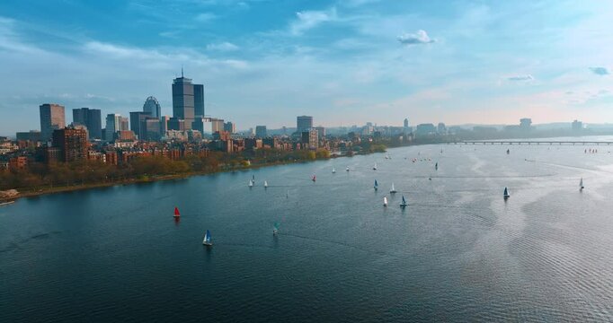 Many boats sailing by the calm waterscape of the Charles River. Drone footage above the waterscape with the view of Boston skyline.