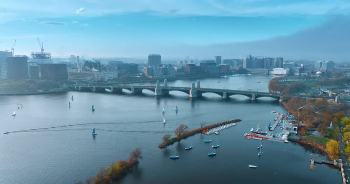 Lots of bots sail by the river Charles in Boston on autumn daytime. Multiple cars move by Harvard Bridge over the river. Aerial view.
