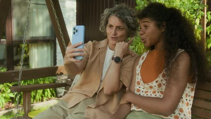 Medium arc shot of joyous Caucasian woman and teenage African American daughter relaxing together on bench swing outdoors, taking selfie on smartphone while smiling and embracing - Powered by Adobe
