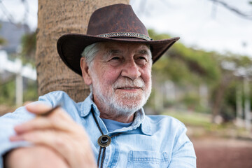 Portrait of a smiling senior Caucasian man with beard sitting outdoors against a tree trunk,...