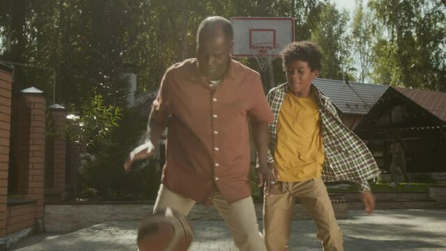Medium full shot of middle-aged African American man playing basketball with teenage son outdoors on sunny summer day, dribbling ball and ducking while boy is trying to tackle and steal it