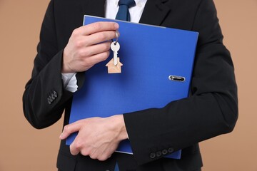 Real estate agent holding key with house shaped keychain and folder on beige background, closeup
