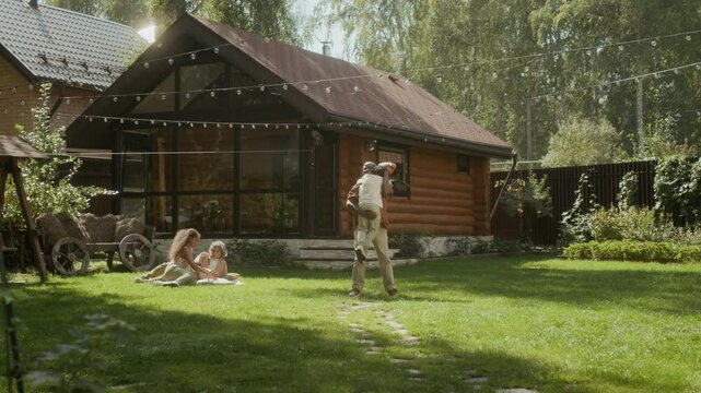 Wide shot of black dad training teenage son in baseball skills on green lawn of suburban family home, then hugging little boy running towards him. Family relaxing together on hot summer day