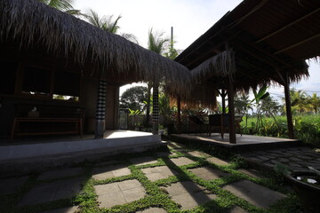 Small house with a porch and a patio. The porch has a bench and a chair. The patio has a table and a couple of chairs. The house is surrounded by trees and has a bamboo roof