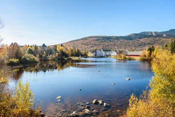 Obraz premium Lake surrounded by forest at the peak of fall foliage in a mountain village on a clear autumn day