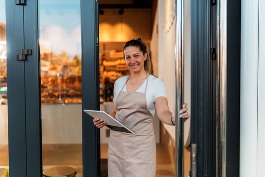 Cheerful baker woman, wearing an apron, opening the bakery door while holding a tablet, warmly inviting customers to enter and indulge in fresh, delicious pastries