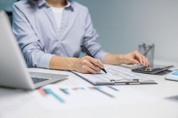 A person diligently works on financial documents at a desk, using a calculator and laptop. The...