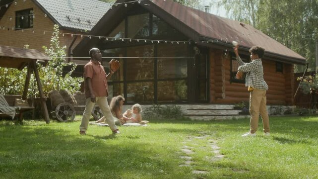 Wide shot of 12-year-old African American boy practicing baseball throwing and catching skills with dad wearing glove, while relaxing outdoors on green lawn with family in bright sunlight haze