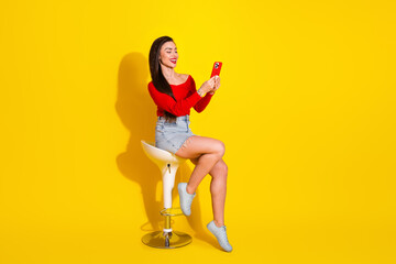 Smiling young woman in a red top taking a photo with a smartphone against a bright yellow background, relaxed and fashionable