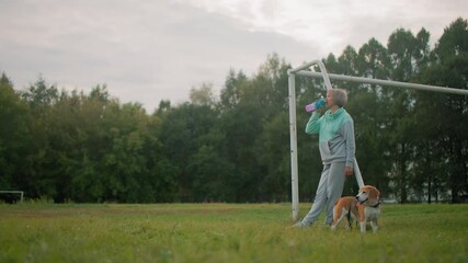 Graceful woman in mint and gray activewear relaxing after morning exercise, drinking from colorful water bottle while standing next to her playful beagle puppy on grassy sports field under cloudy sky - Powered by Adobe