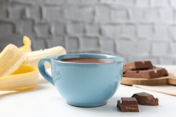 Tasty chocolate milk in cup, pieces and banana on white wooden table, closeup