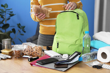 Survival kit. Woman packing different emergency supplies into backpack at wooden table indoors,...
