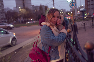 Fototapeta premium Woman waiting at a street corner in the early evening with traffic and city lights in the background