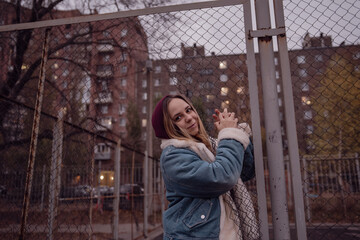 A young woman in a cozy jacket leans against a chain link fence at an urban park during autumn in...
