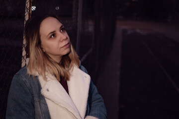Fototapeta premium A young woman stands near a chain-link fence in an urban area during the late afternoon of an autumn day