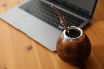 Traditional yerba mate tea and laptop on wooden table, closeup