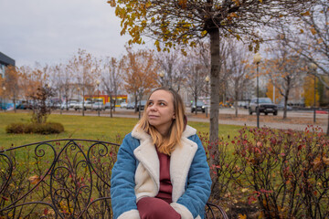 A woman sitting thoughtfully on a park bench surrounded by autumn foliage in an urban setting...