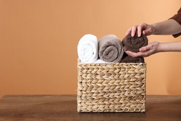 Woman taking clean towel from basket at wooden table against brown background, closeup. Space for...