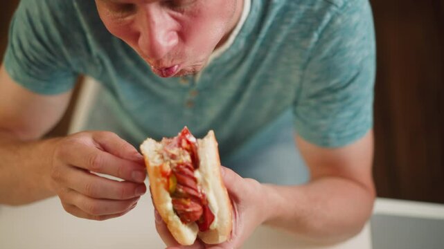 Close up of hungry boy biting into juicy burger for dinner, holding sandwich with both hands over white table, wearing casual teal shirt, filled with sausage, tomato, sauce and peper