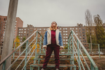 A confident woman stands on rusted stairs in a dilapidated urban area during a cloudy autumn day