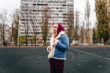 A young woman in a denim jacket and hoodie stands thoughtfully in front of an urban building on a...