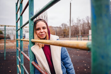 Obraz premium Young woman in a hoodie and coat poses at a playground during autumn in a city park, surrounded by colorful trees and apartment buildings