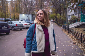 Fototapeta premium A woman stands thoughtfully in an urban setting during autumn, surrounded by trees and parked cars in a quiet neighborhood