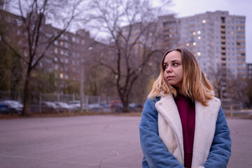 Fototapeta premium Young woman in casual attire stands in an urban park during early evening, surrounded by tall buildings and autumn trees