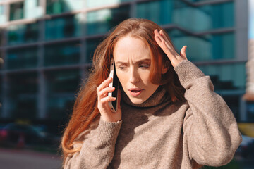 Fototapeta premium Woman with long red hair talking on phone in urban setting, expressing concern while wearing a cozy sweater during daylight hours