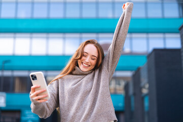 Woman celebrating with joy outdoors while looking at her smartphone in an urban setting during...