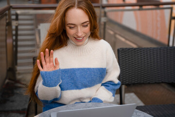 Woman in blue and white sweater waves while working on laptop at outdoor café during daytime,...