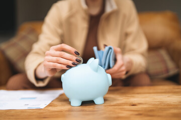 Person placing money into a light blue piggy bank while sitting on a couch in a cozy living room...