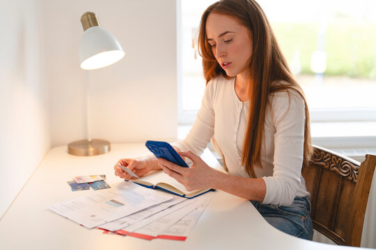 Young woman managing personal finances while using smartphone at home desk in morning light