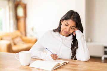 Young woman writing in a notebook while sitting at a wooden table in a cozy living room during the day