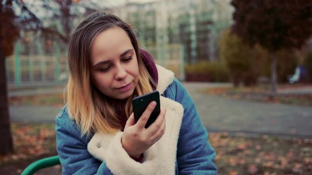 A woman dressed in a warm blue jacket sits on a bench in a city park, focused on her phone as autumn leaves fall around her on a cloudy day.