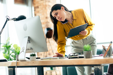 Focused businesswoman multitasking in a stylish office workspace with a computer and clipboard on a...