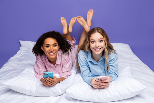 Two young women in pajamas lying on a bed using smartphones on a violet background, sharing good moments together - Powered by Adobe
