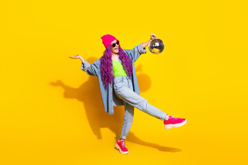 Trendy woman with bright pink hair and stylish outfit posing playfully against a vibrant yellow background, holding a shiny disco ball