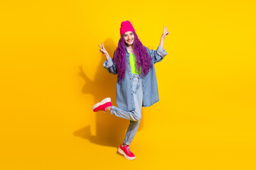 Trendy young woman in denim and bright outfit making peace signs against yellow background...
