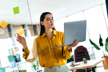 Zelfklevend Fotobehang Young businesswoman strategizing office tasks with post it notes and laptop in a modern and bright workspace interior © deagreez