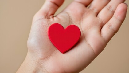 Obraz premium A hand holds a red heart-shaped object, symbolizing love and affection against a soft, neutral background.