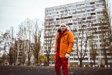 Man in orange jacket stands in urban park surrounded by bare trees and large apartment building...