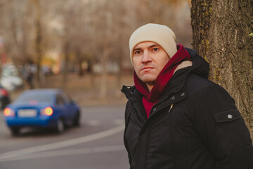 Fototapeta premium Man in winter attire stands by a tree on a city street during autumn, contemplating the passing vehicles