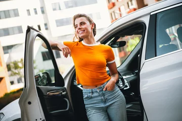 Fotobehang Young cheerful woman standing next to a car door in a vibrant urban setting during daylight, radiating confidence and charm © deagreez