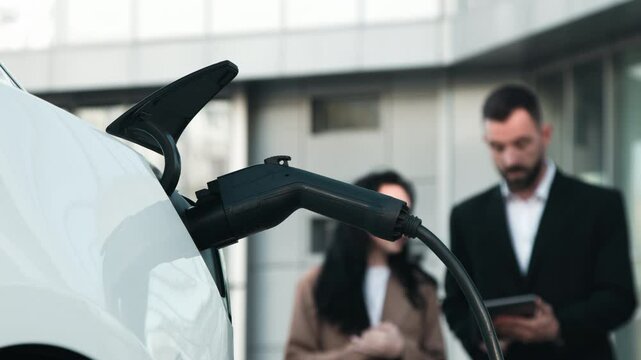 Charging an electric car in the background of two business people discussing the project. A man takes the car off the charger. Concept of ecological transportation and business.