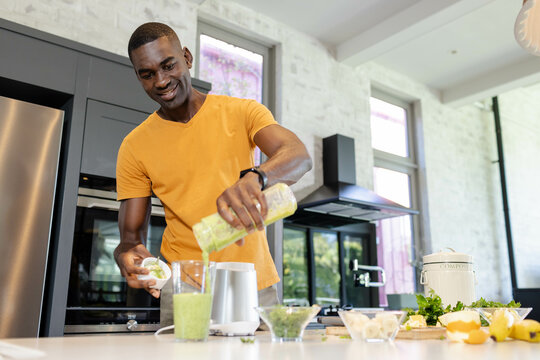 African American man pouring vibrant green smoothie in home kitchen, with blender jar, compost bin - Powered by Adobe